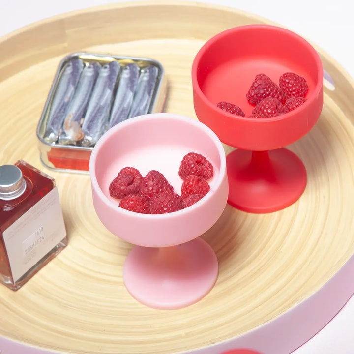 Two small silicone cocktail glasses with raspberries on a wooden surface, next to a small bottle and container.