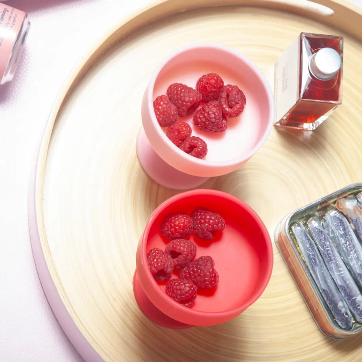 Two pink silicone cocktail glasses filled with raspberries on a wooden surface.