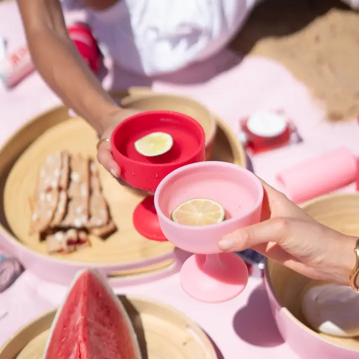 Person holding a pink silicone cocktail glass with water and a slice of lime, with various dishes and a watermelon.