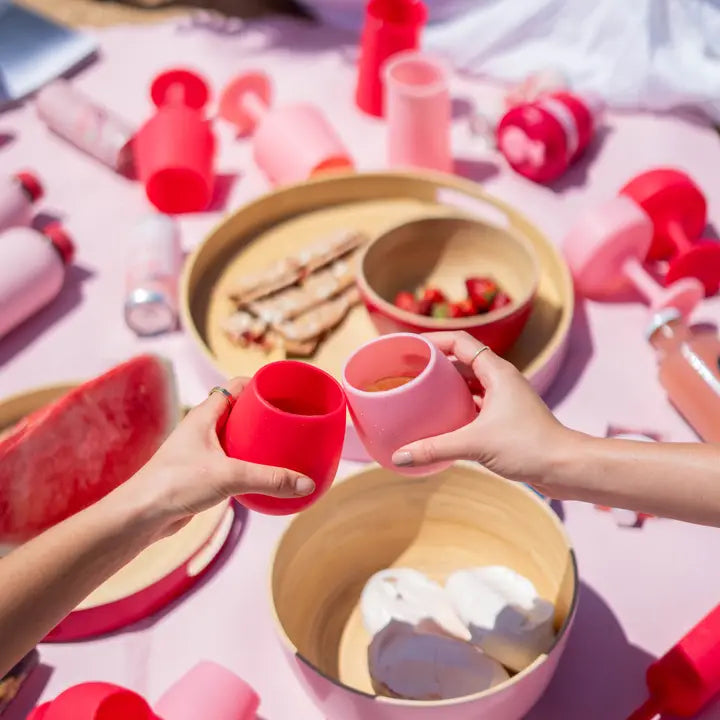 Two hands holding a red and a pink silicone cup toasting together on a pink picnic blanket surrounded by matching pink and red silicone drinkware and bamboo bowls.
