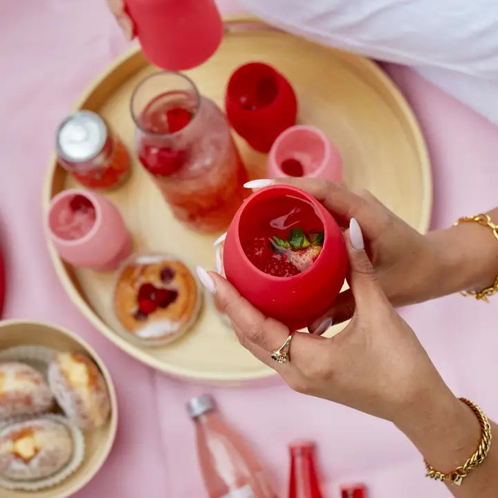 A person holds a bright pink silicone cup filled with a strawberry drink, with matching pink cups and bottles arranged on a round wooden tray on a pink surface.