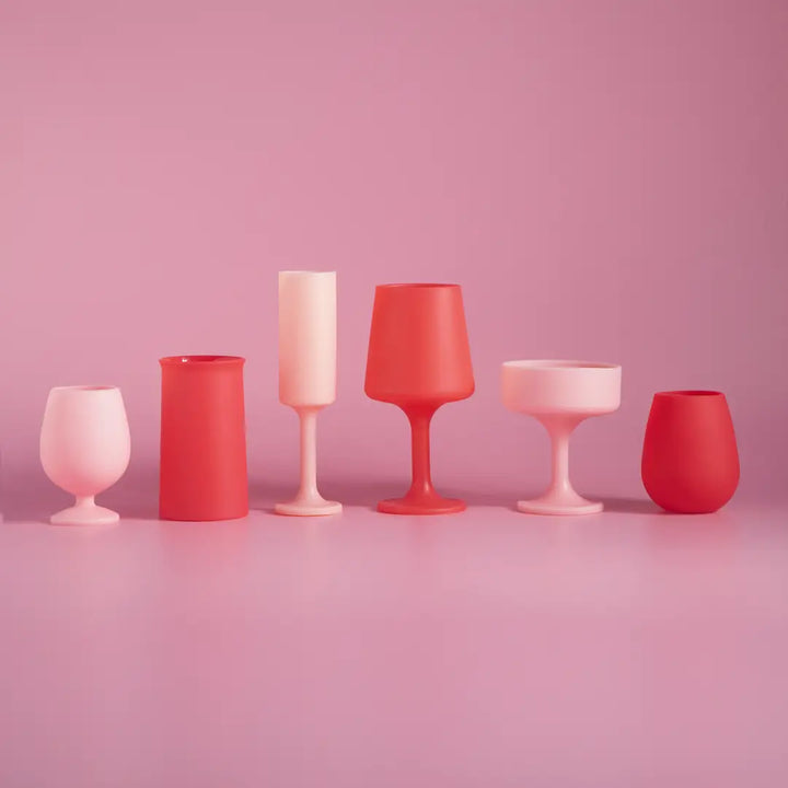 Six matte silicone glassware pieces in soft pink and coral-red tones arranged in a row against a solid pink studio background.