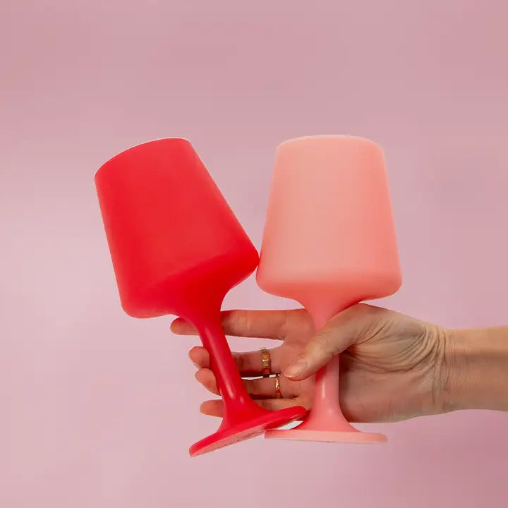 Two silicone unbreakable wine glasses, one red and one light pink, held by a hand against a solid pink background.