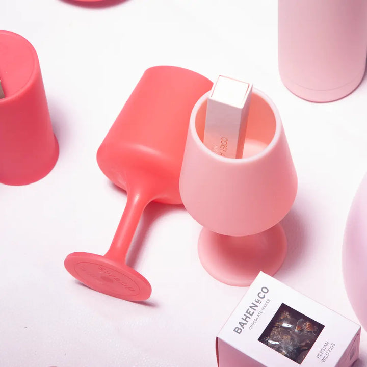 Pink lip balm in a pink silicone wine glass with a red wine glass laying down on a light pink background.