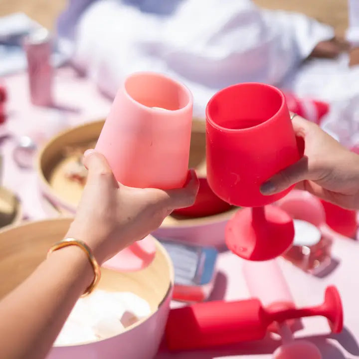 Two hands holding pink silicone wine glasses in a bright, pink-toned setting with other matching pink items blurred in the background.