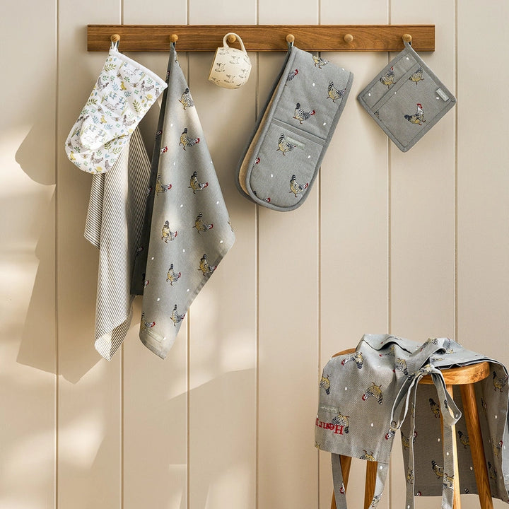 A gray kitchen tea towel featuring a repeating pattern of chickens and small white dots, shown flat against a white background, hanging on a rack that also includes a matching pot holder, double oven glove and apron