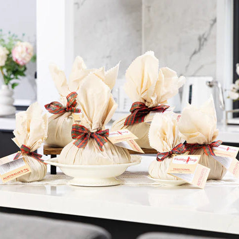 Five wrapped Christmas puddings tied with calico and red ribbon and attached paper tags, arranged on a white countertop in a bright kitchen setting.