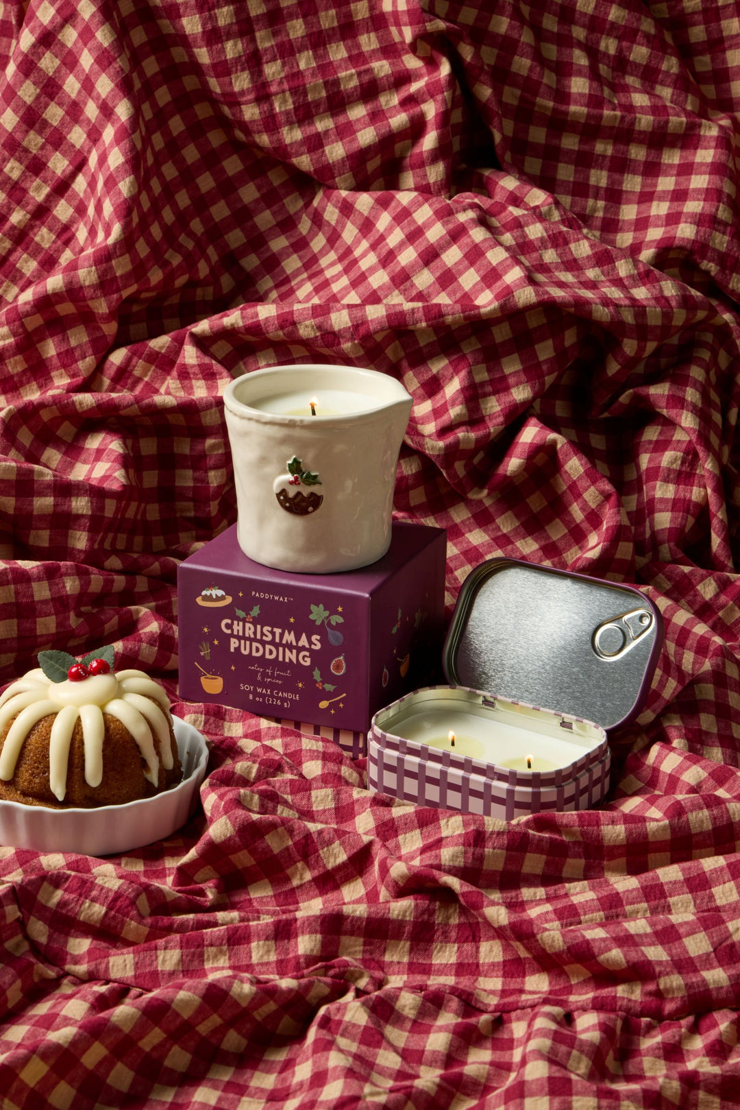 A styled product shot of a candle set on red and beige checkered fabric. The set includes a white ceramic candle in a dark red pot with a pudding illustration, its purple &