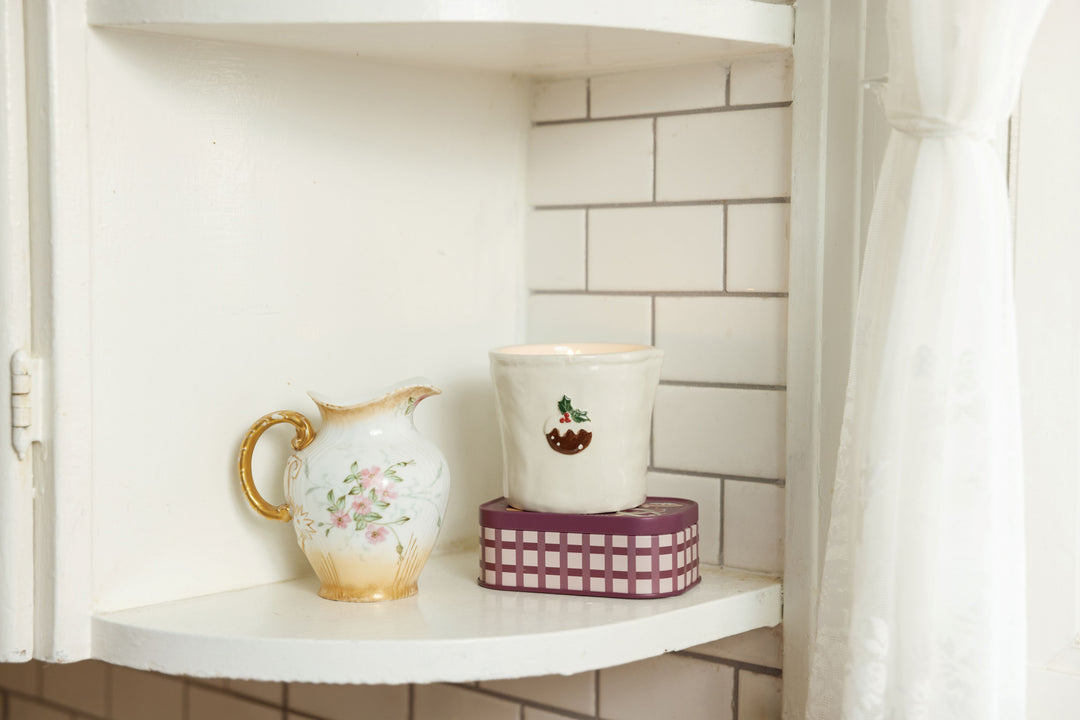 A white ceramic pitcher with gold trim and pink floral detailing sits on a white shelf next to a white cup with Christmas pudding design, stacked on top of a purple-and-white checkered tin, against a white subway tile wall.