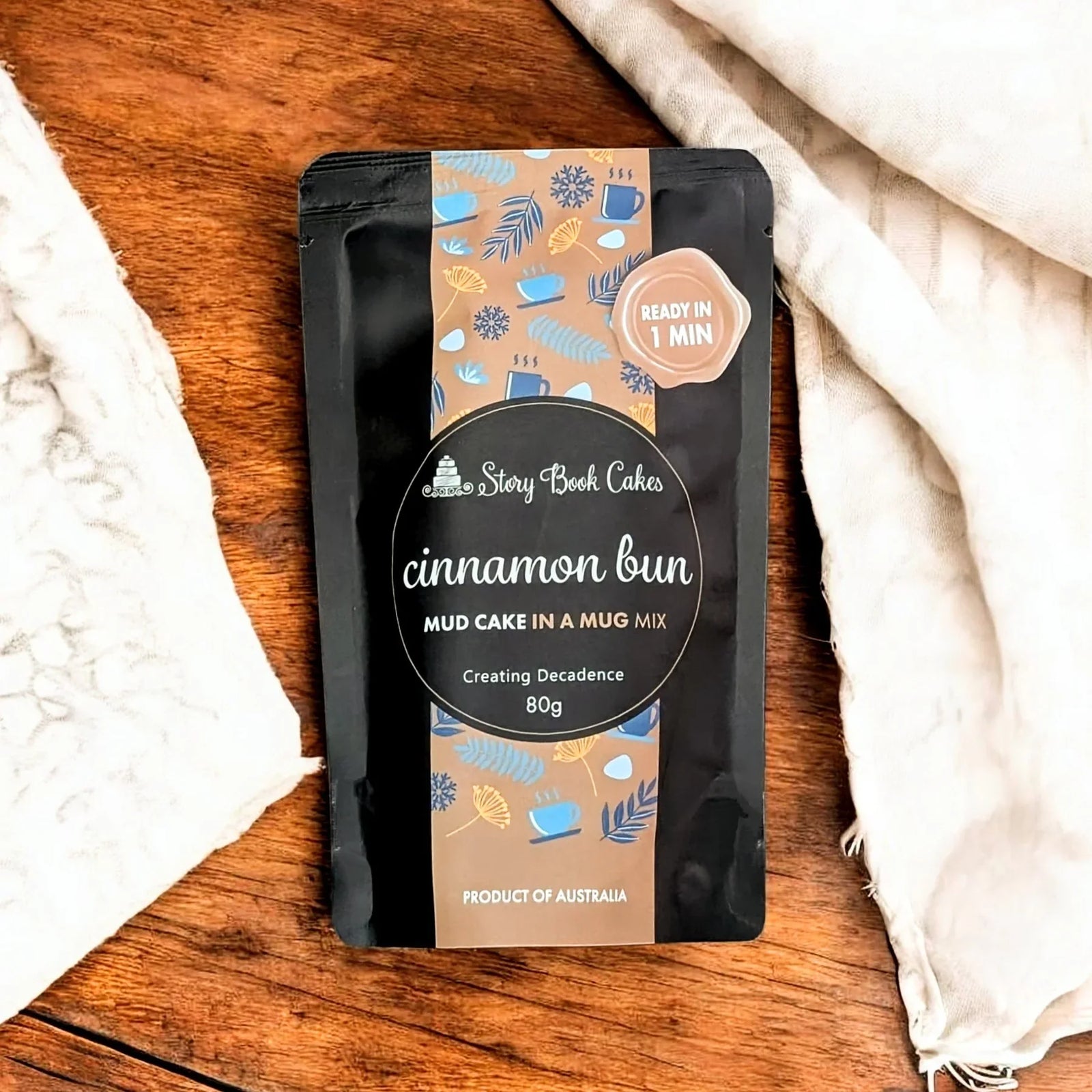 Packaged cinnamon bun mud cake in a mug mix on a wooden surface with a white cloth.