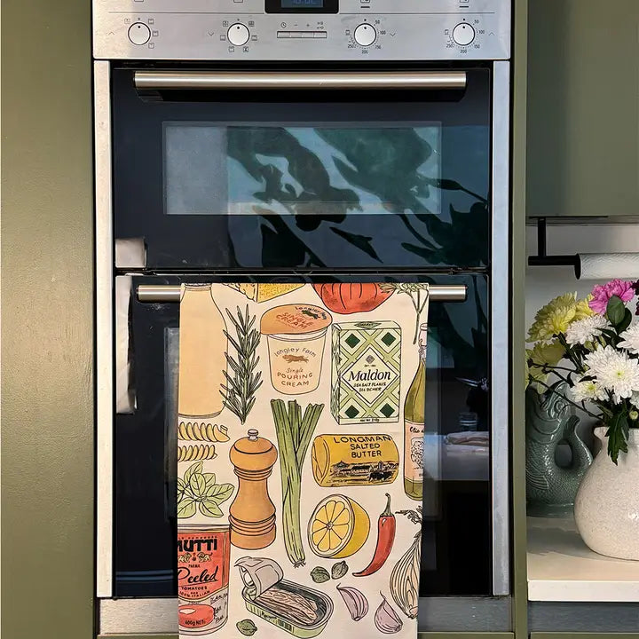 A kitchen towel with a colorful illustrated print of food items like pasta, cheese, garlic, and vegetables, hanging from the metal handle of a built-in oven in a kitchen.