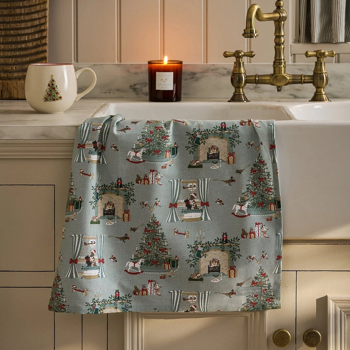 A rectangular fabric tea towel draped over a sink, featuring a festive Christmas pattern on a light blue-gray background. The pattern includes Christmas trees, stockings, fireplaces with Santa Claus, and small birds.