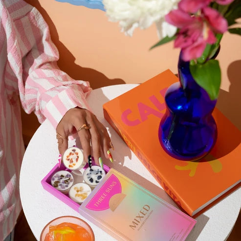 A styled flat-lay featuring a pink and white striped shirt sleeve holding a pink tray of tealight candles and an orange book with a blue vase of flowers on a white table.