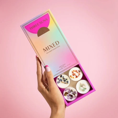 A hand holds an open iridescent pink box labeled THREE SUNS MIXED TEALIGHT COLLECTION against a solid pink background. The box contains three small round cream or wax melts with visible botanical and fruit pieces.