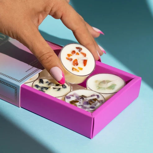 A hand with pink nails holds a small round tin above an open bright pink box containing several similar round tins filled with tealight candles and decorated with dried botanicals, set against a blue background.