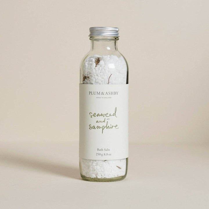 A glass bottle filled with white bath salts and dried botanicals, featuring a white label that reads PLUM &amp; ASHBY seaweed and samphire bath salts 250g, set against a solid beige background.