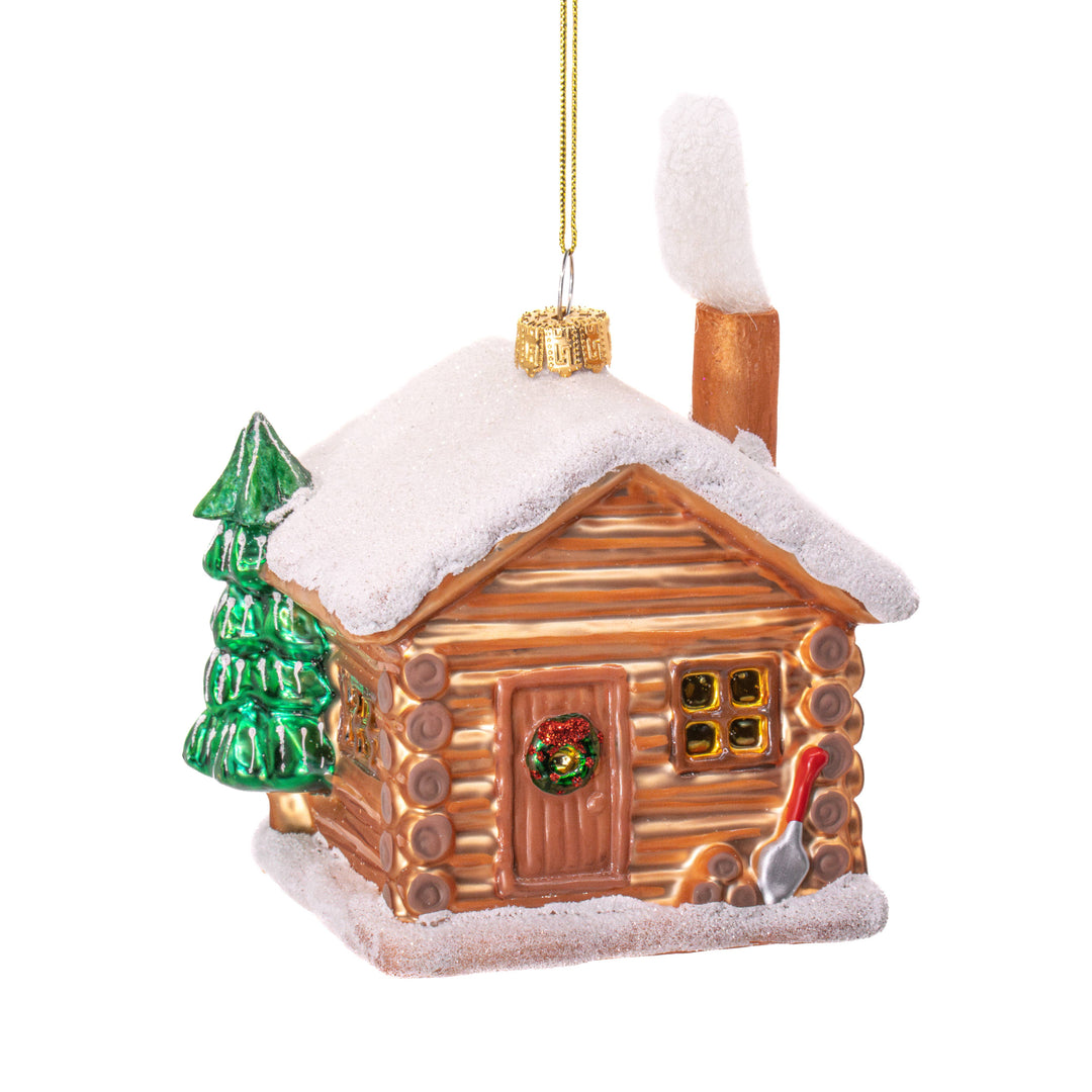 A Christmas ornament shaped like a rustic log cabin with a snow-covered roof, green tree, gold hanging string, and small red-and-white snow shovel detail, shown on a plain white background.