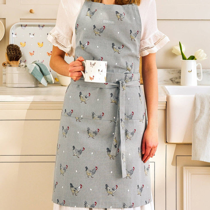 A woman wearing a light gray apron with a colorful chicken and white dot pattern, holding a white mug with a chicken illustration, in a bright kitchen setting.