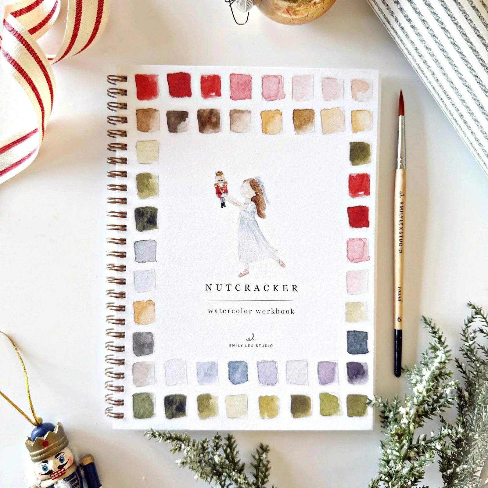 Watercolor workbook titled 'Nutcracker' with a pen and Christmas decorations on a white surface