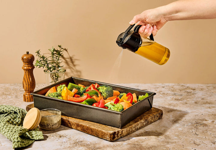 A hand using a dreamfarm oil bottle with a mist of oil spraying out over vegetables in a baking dish
