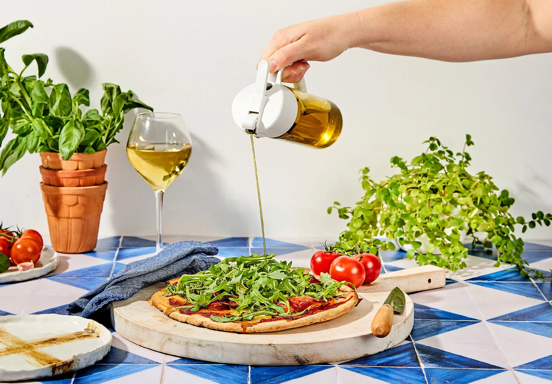 A hand drizzling olive oil from a glass bottle over a pizza