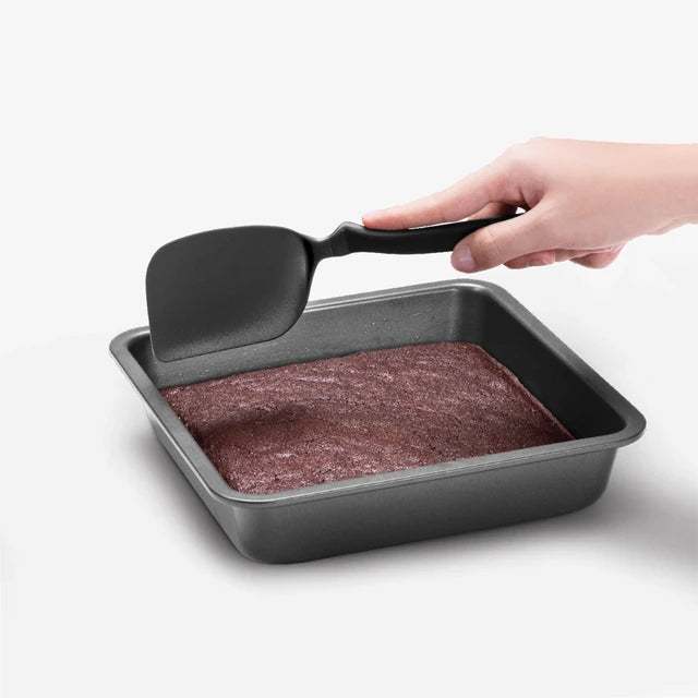 Hand using a spatula to lift a square cake from a baking pan on a white background