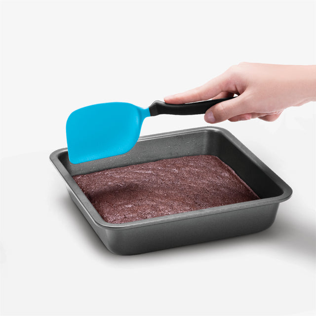 Hand using a blue spatula to lift brownies from a baking pan on a white background