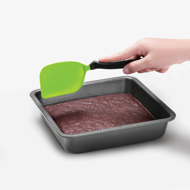 Hand using a green spatula to lift brownies from a baking pan on a white background