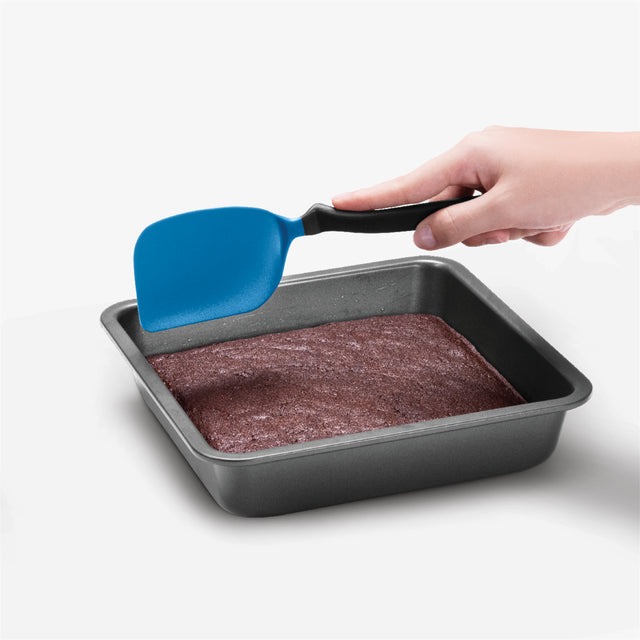 Hand using a blue spatula to lift brownies from a baking pan on a white background