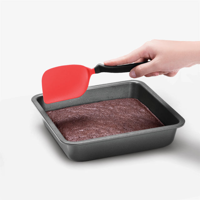 Person using a red spatula to lift brownies from a baking pan on a white background