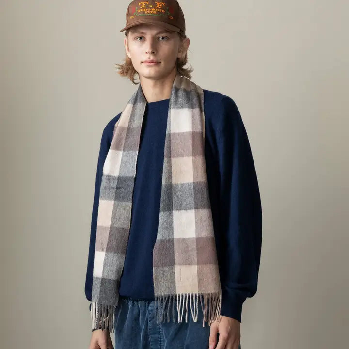 Person wearing a plaid scarf, navy sweater, and brown cap against a neutral background. 