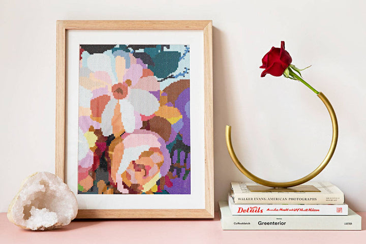 A framed abstract floral sparkle art artwork in a light wood frame, styled on a pink surface with a white crystal, books, and a gold rose holder against a white wall.