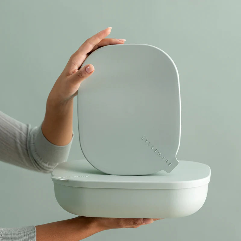 A person holds a light gray silicone lid with the brand name 'STYLEWARE' embossed on it, resting on top of a matching light gray oval container against a pale green background.