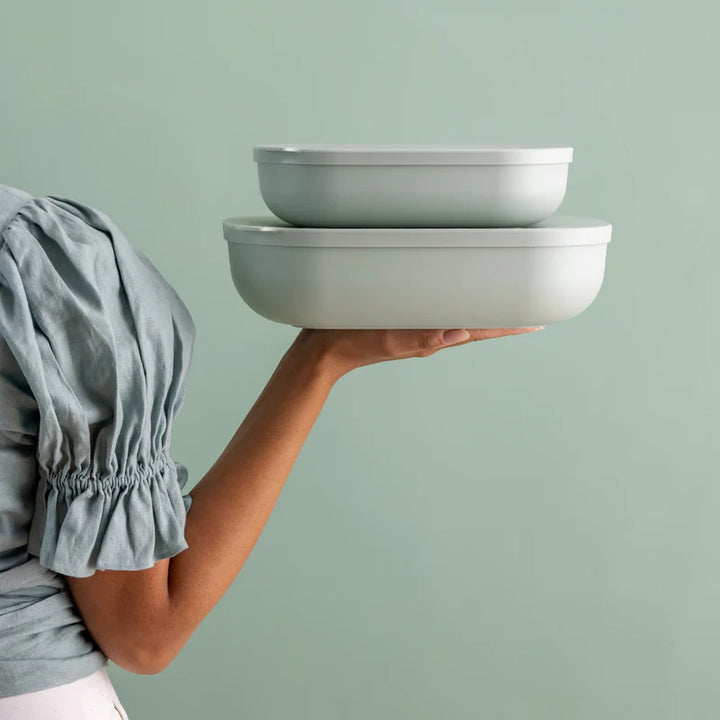 A person in a gray puff-sleeve top holds two stacked light green oval containers against a solid pale green background.