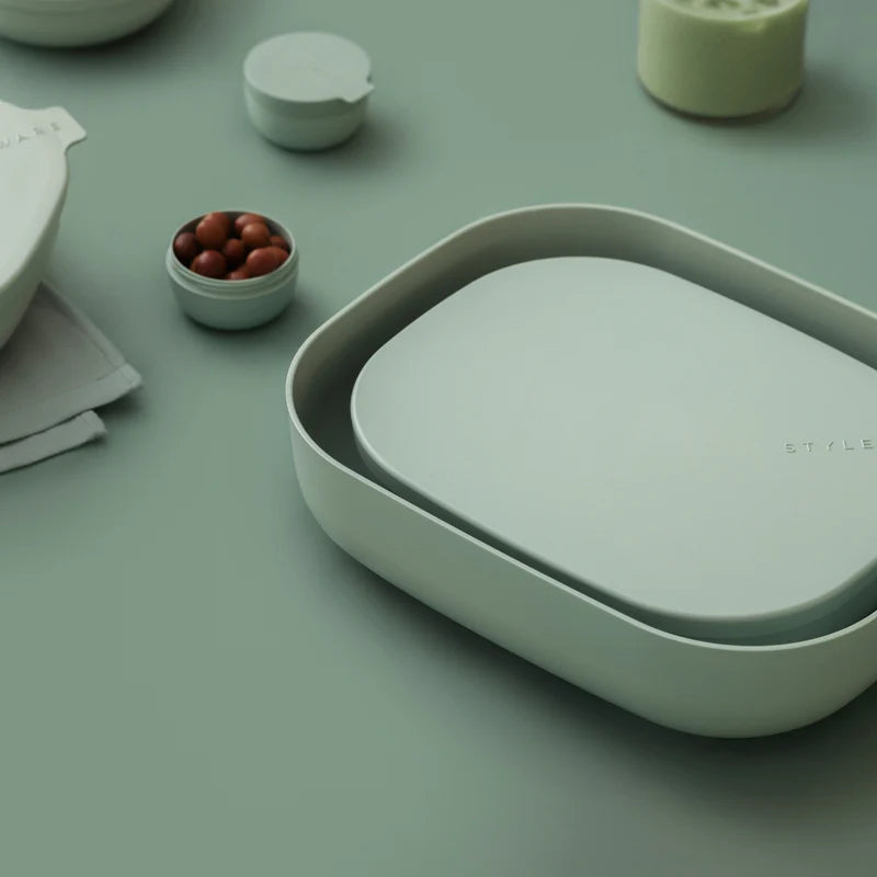 A minimalist light sage green oval-shaped container with a matching lid, styled on a matching sage green surface with other abstract home accessories in the background.