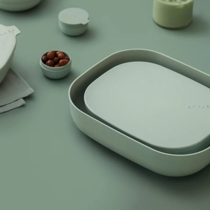 A minimalist light sage green oval-shaped container with a matching lid, styled on a matching sage green surface with other abstract home accessories in the background.