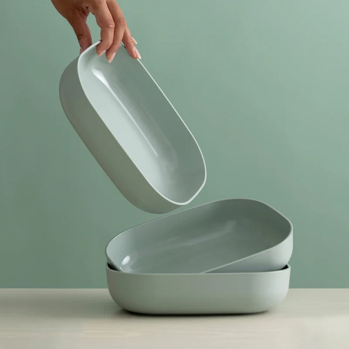 A hand holds a light sage green oval tray above two stacked matching trays on a light surface against a soft sage green background.