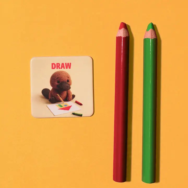 A square card with the word "DRAW" and an illustration of a platypus drawing, placed beside two colored pencils on a solid yellow background.