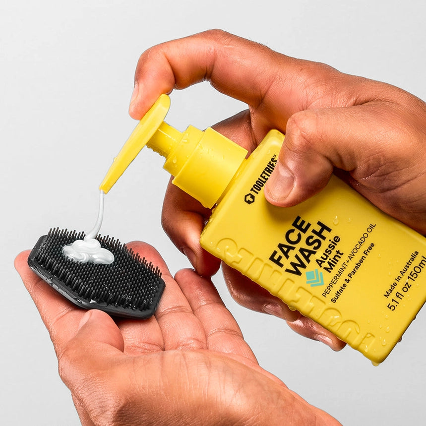 Hands pumping face wash from a yellow bottle onto a black facial exfoliating scrubber, in front of a white background