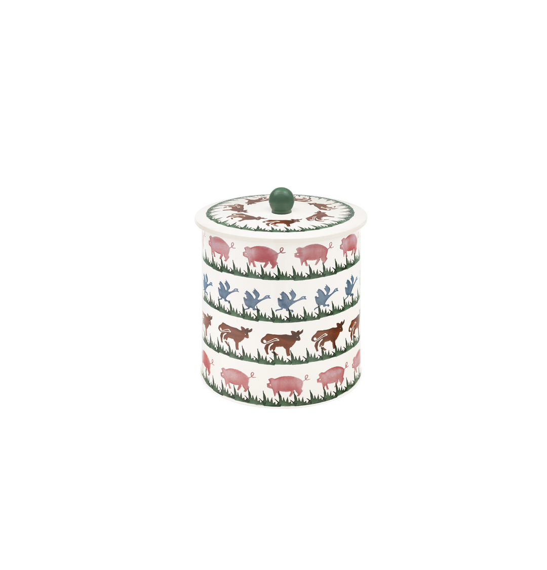 A cylindrical lidded metalcontainer with a white background and a repeating pattern of pink pigs, green grass, and blue birds &amp; brown cows topped with a small green knob.