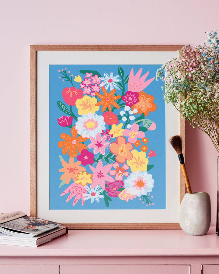 Framed colorful floral artwork on a pink wall with a vase and books below.