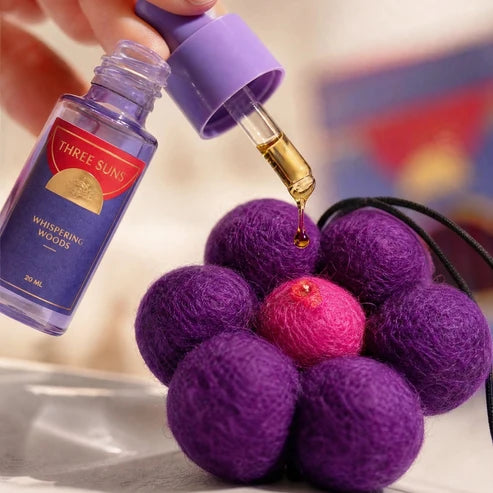 A hand holds a purple dropper bottle of Three Suns fragrance oil above a purple felt flower-shaped ornament with a bright pink center, with a drop of golden oil being dripped onto it.