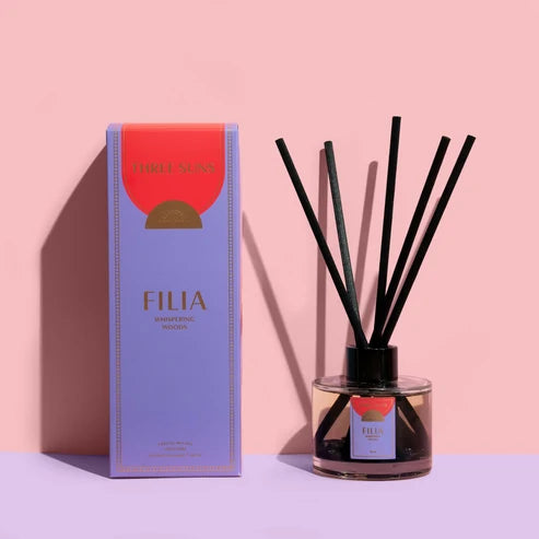A reed diffuser bottle with black reeds next to its purple and orange packaging box, set against a solid pink background.