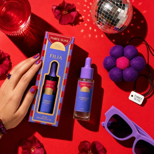 A hand holds a colorful product box next to a small dropper bottle labeled FILIA by Three Suns, arranged on a bright red surface with flowers, sunglasses, a disco ball, and purple accessories.
