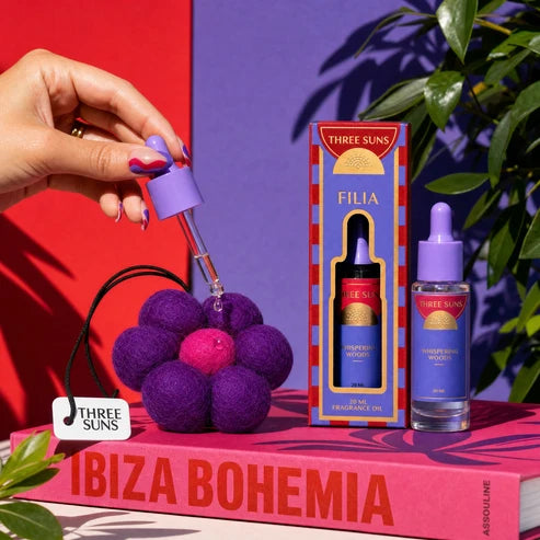 Three Suns Filia set, featuring a hand holding a purple dropper over a purple felt flower, with the retail box and glass bottle standing on a pink book against a red and purple background.
