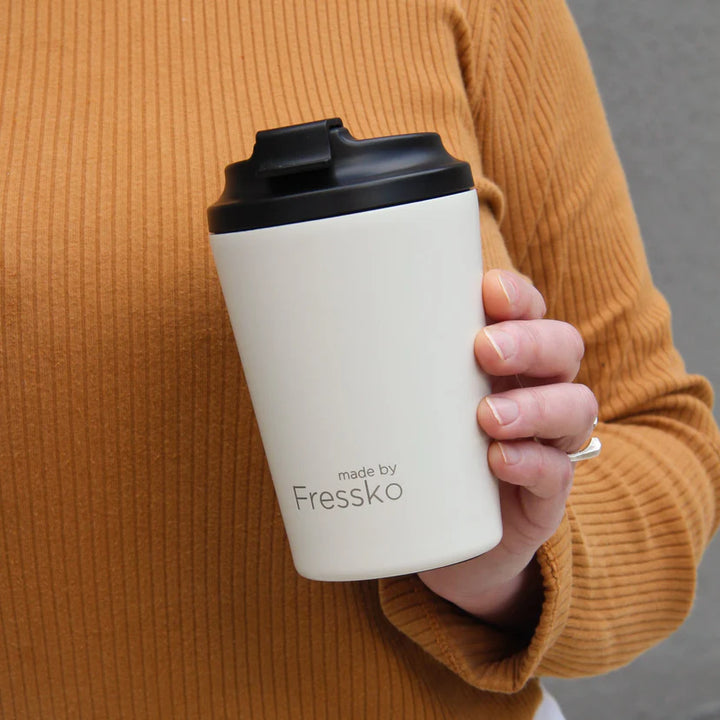 Person holding a white coffee cup with black lid, branded &