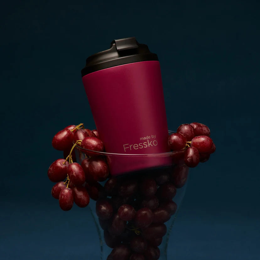 Maroon travel cup with black lid surrounded by red grapes on a dark background
