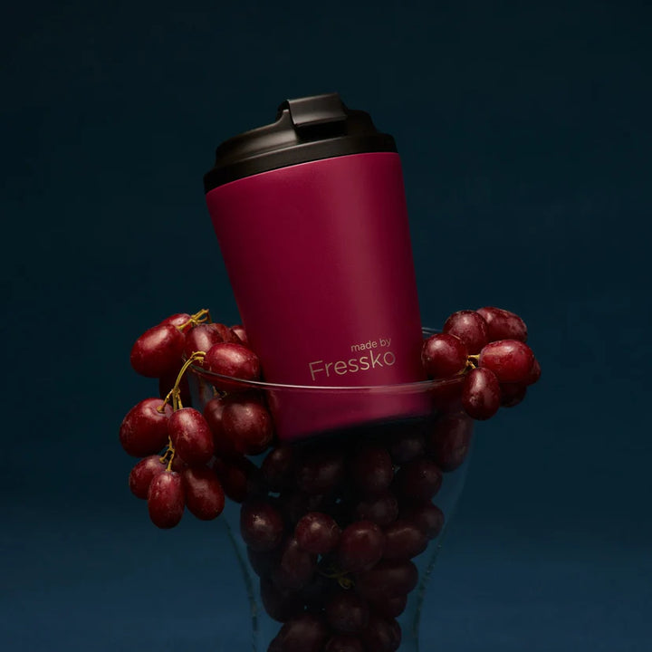 Maroon travel cup with black lid surrounded by red grapes on a dark background