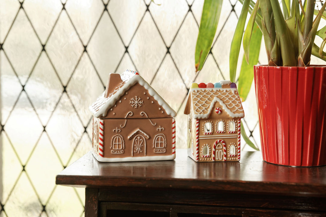 Two gingerbread house ceramic ornaments displayed on a dark wooden table next to a red potted plant, with a diamond-patterned window in the background.
