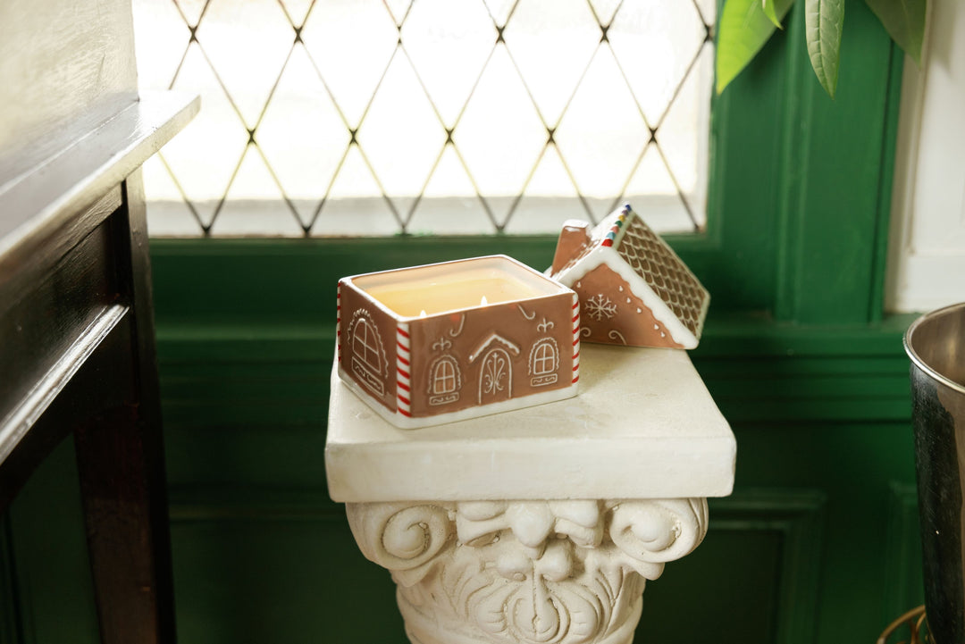 A gingerbread-themed candle in a ceramic container with a house design, displayed on a white stone pedestal in front of a green paneled wall and diamond-patterned window